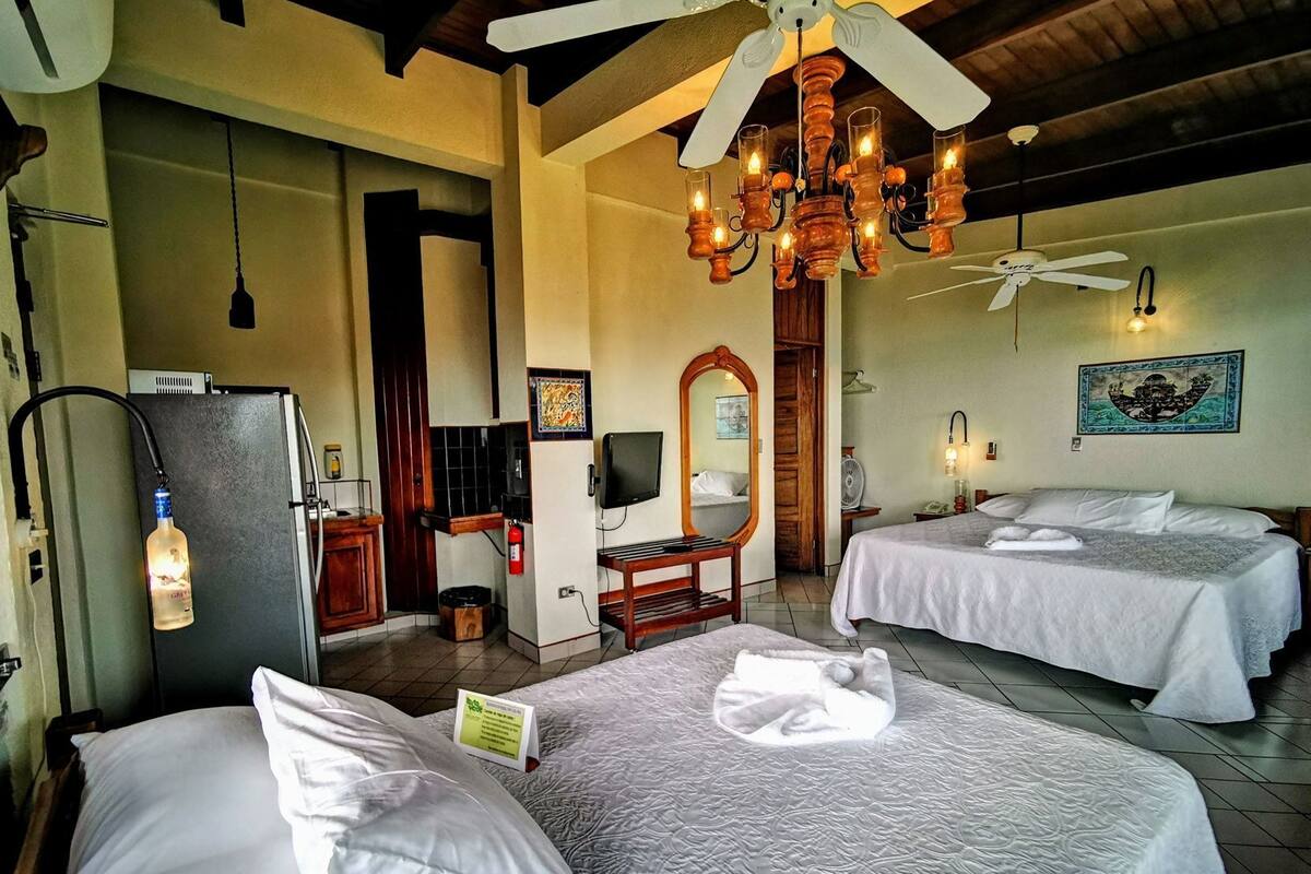 A hotel room with two beds, a ceiling fan, a chandelier, a refrigerator, a television, and a painting on the wall.
