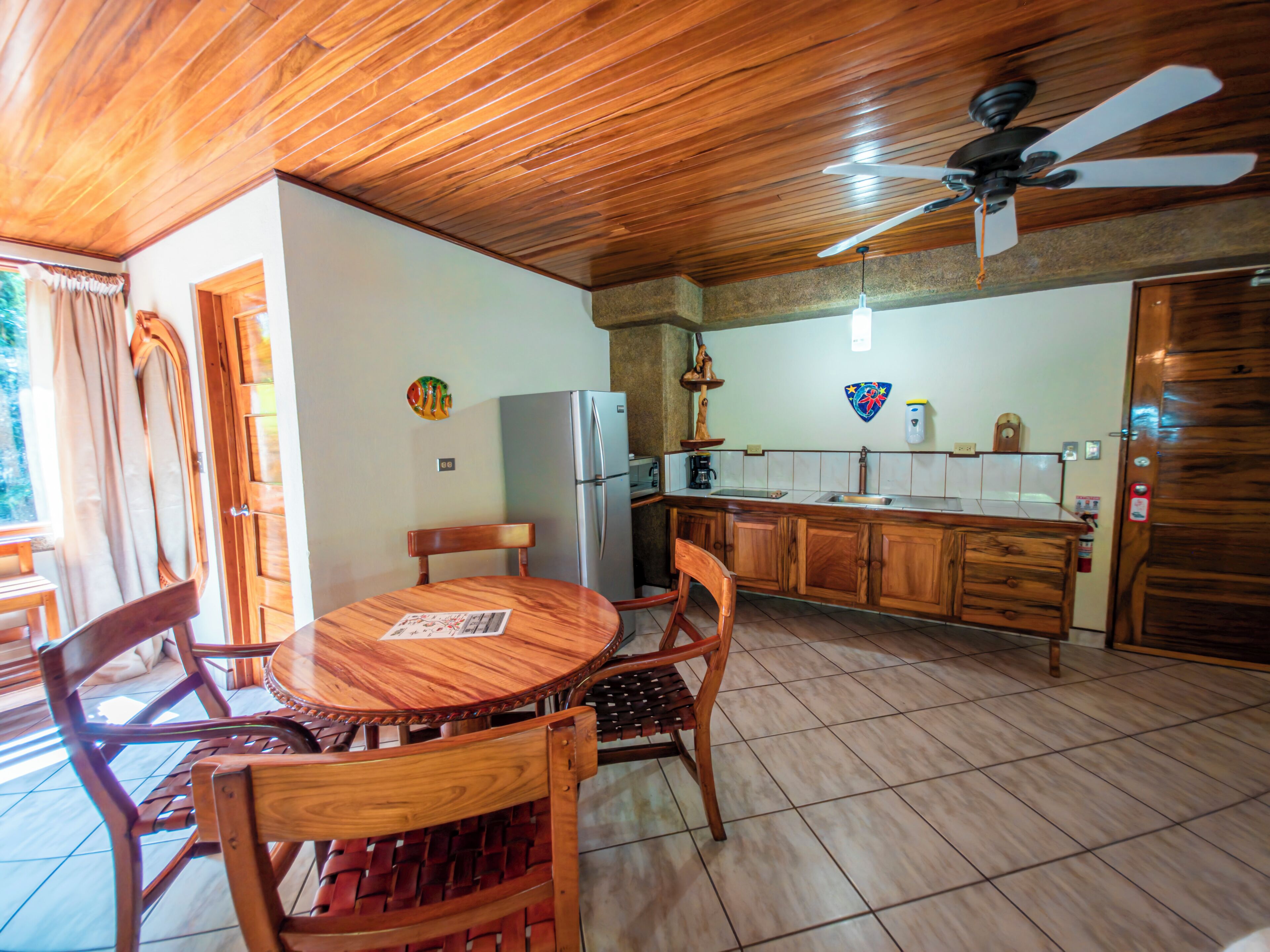 Ein Holzespeisetisch mit Stühlen, eine Küche inklusive Kühlschrank und ein Deckenventilator.