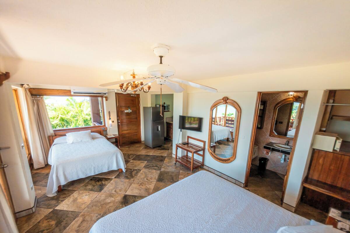 A hotel room with two beds, a ceiling fan, a TV, a mirror, and a view of the outdoors.