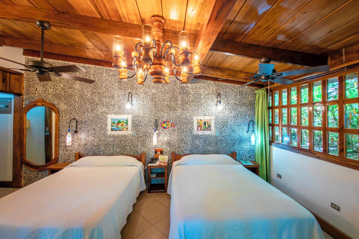A room with two beds, a chandelier, a ceiling fan, and a window with a view of greenery.