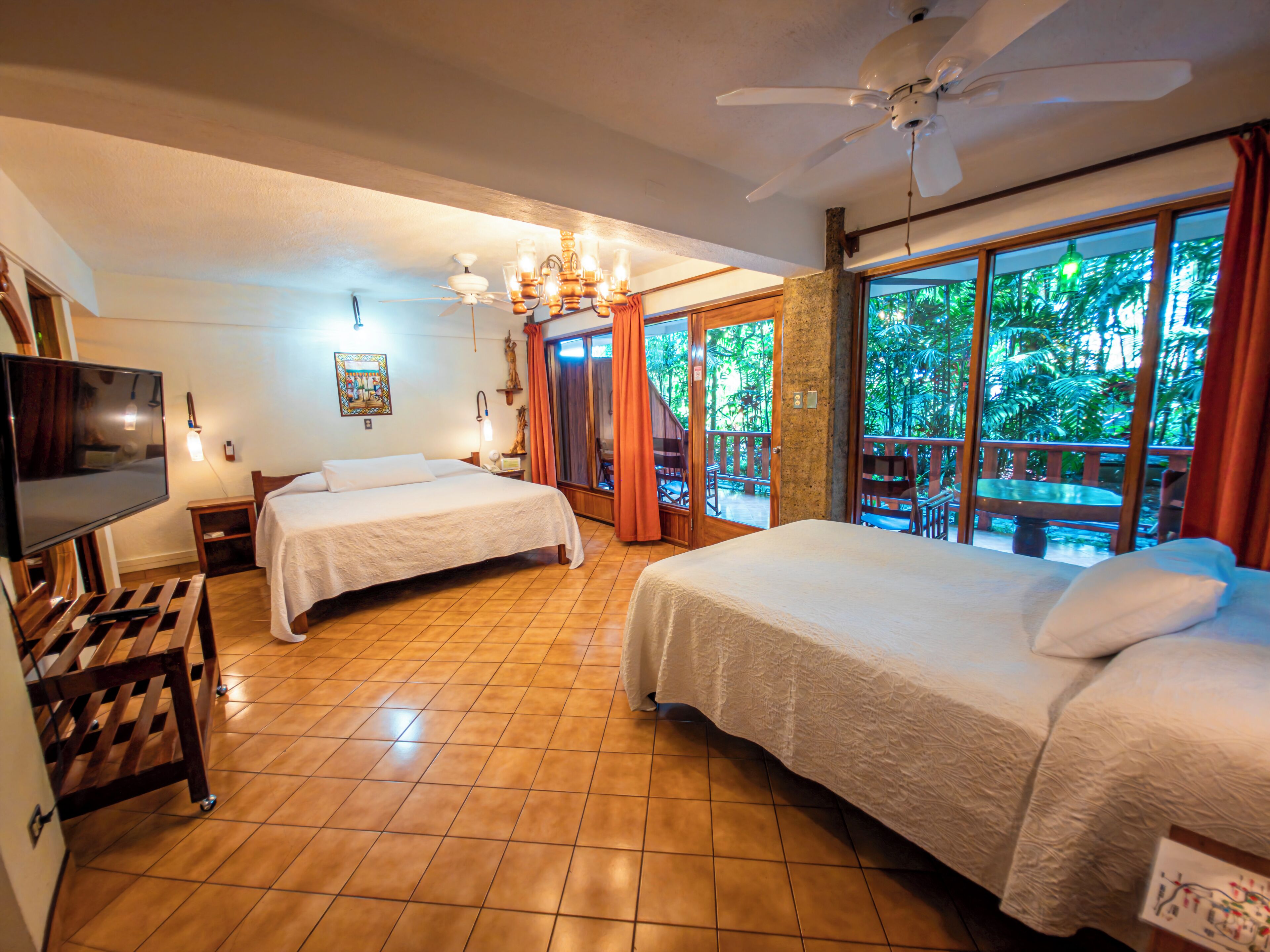 Habitación de hotel con dos camas, televisión, ventilador de techo y balcón con vistas a zonas verdes.