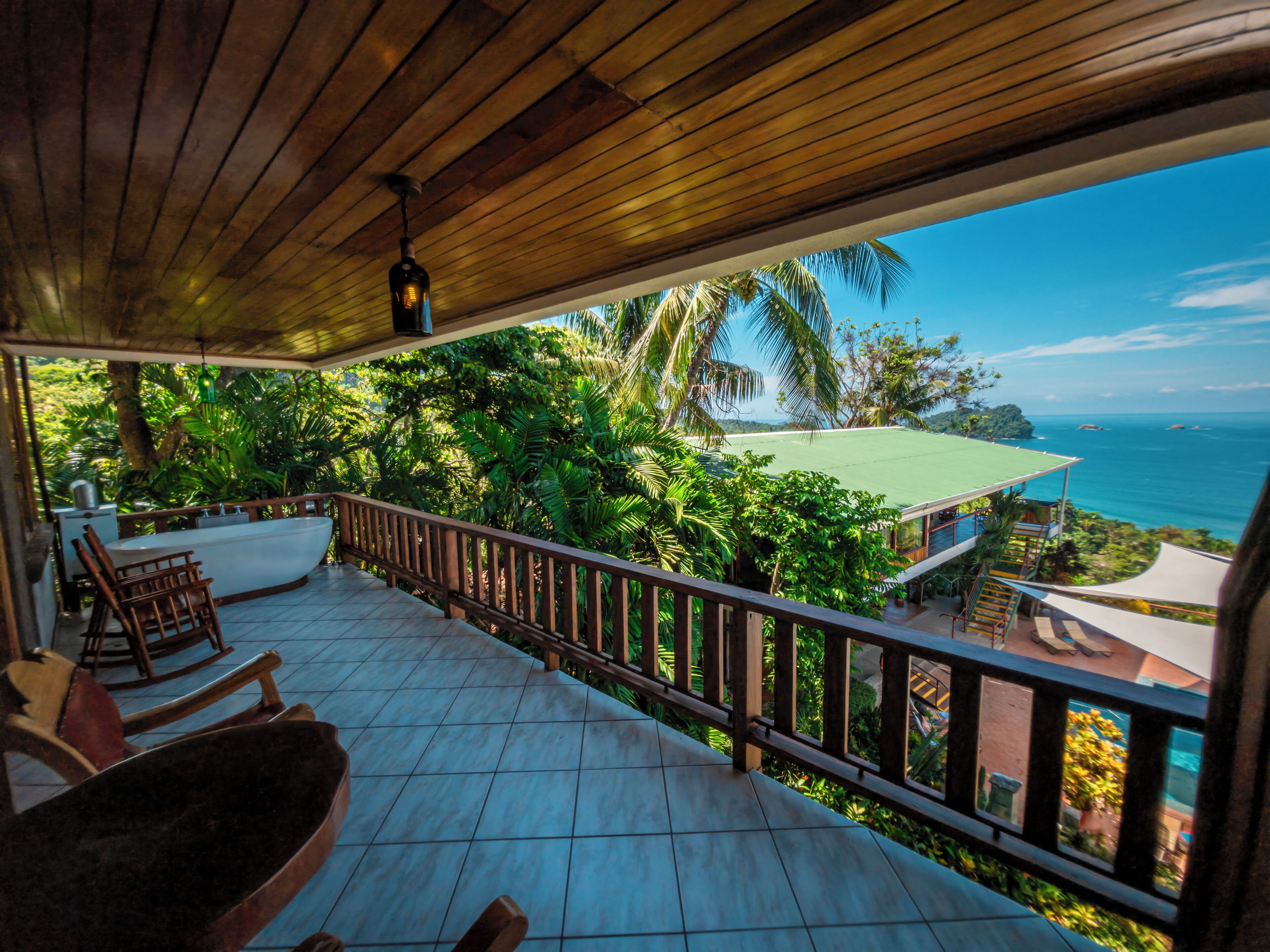 A covered balcony with a view of the ocean, a pool, and lush greenery.
