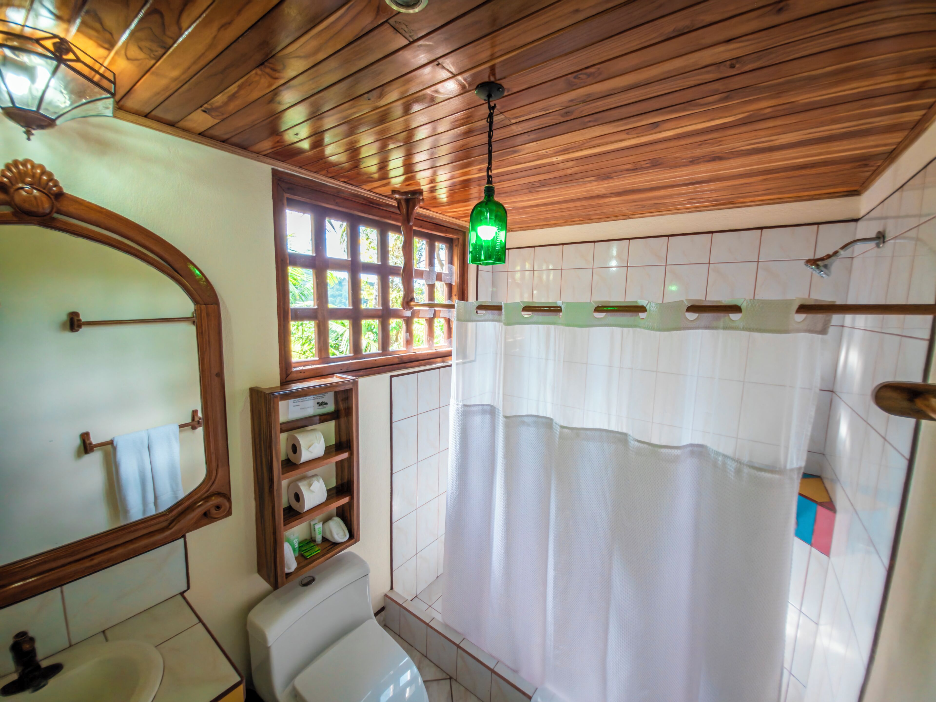A bathroom with a wooden ceiling, a toilet, a sink, a mirror, a shower curtain, and a window with a view.