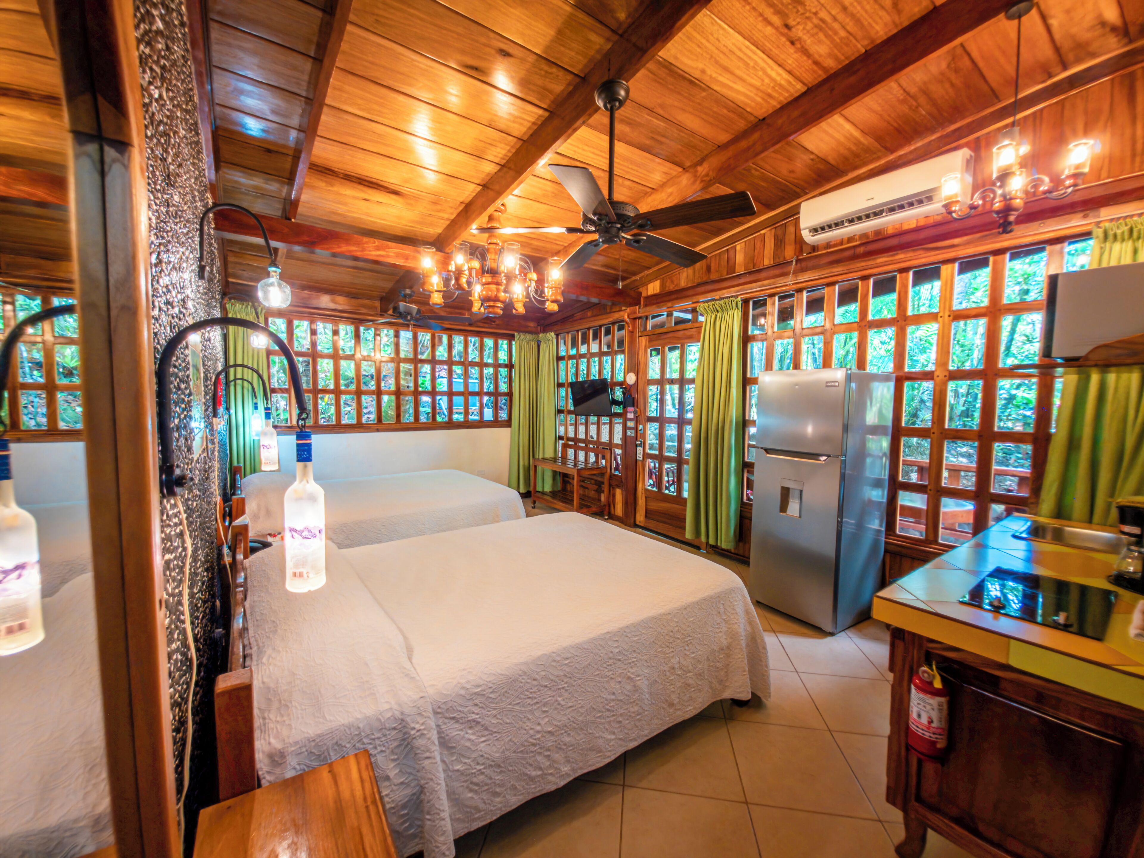 A wooden cabin with a bed, a ceiling fan, a refrigerator, and a window with a view of greenery.
