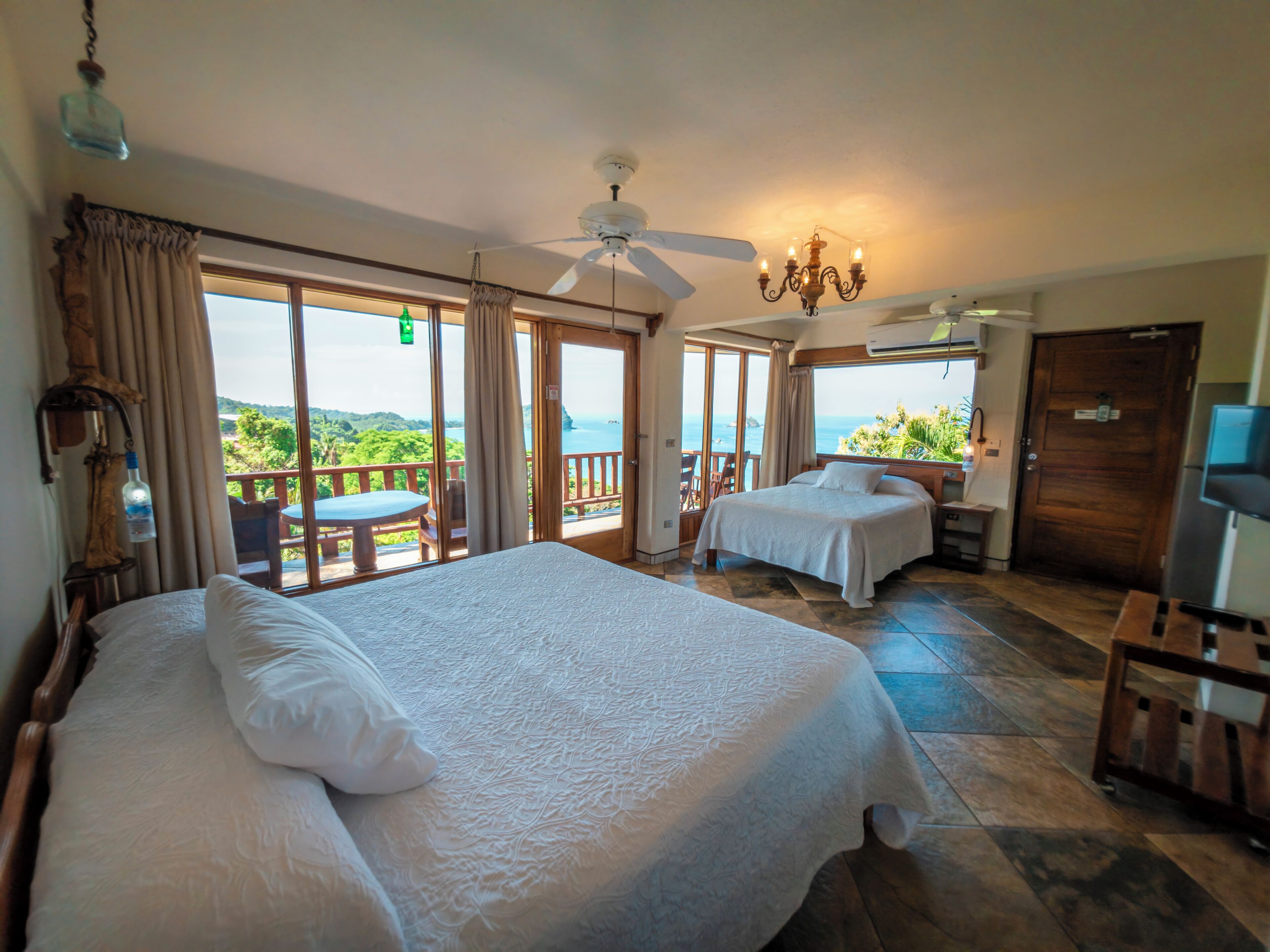 A hotel room with a large bed, a smaller bed, a ceiling fan, a chandelier, and a view of the sea.