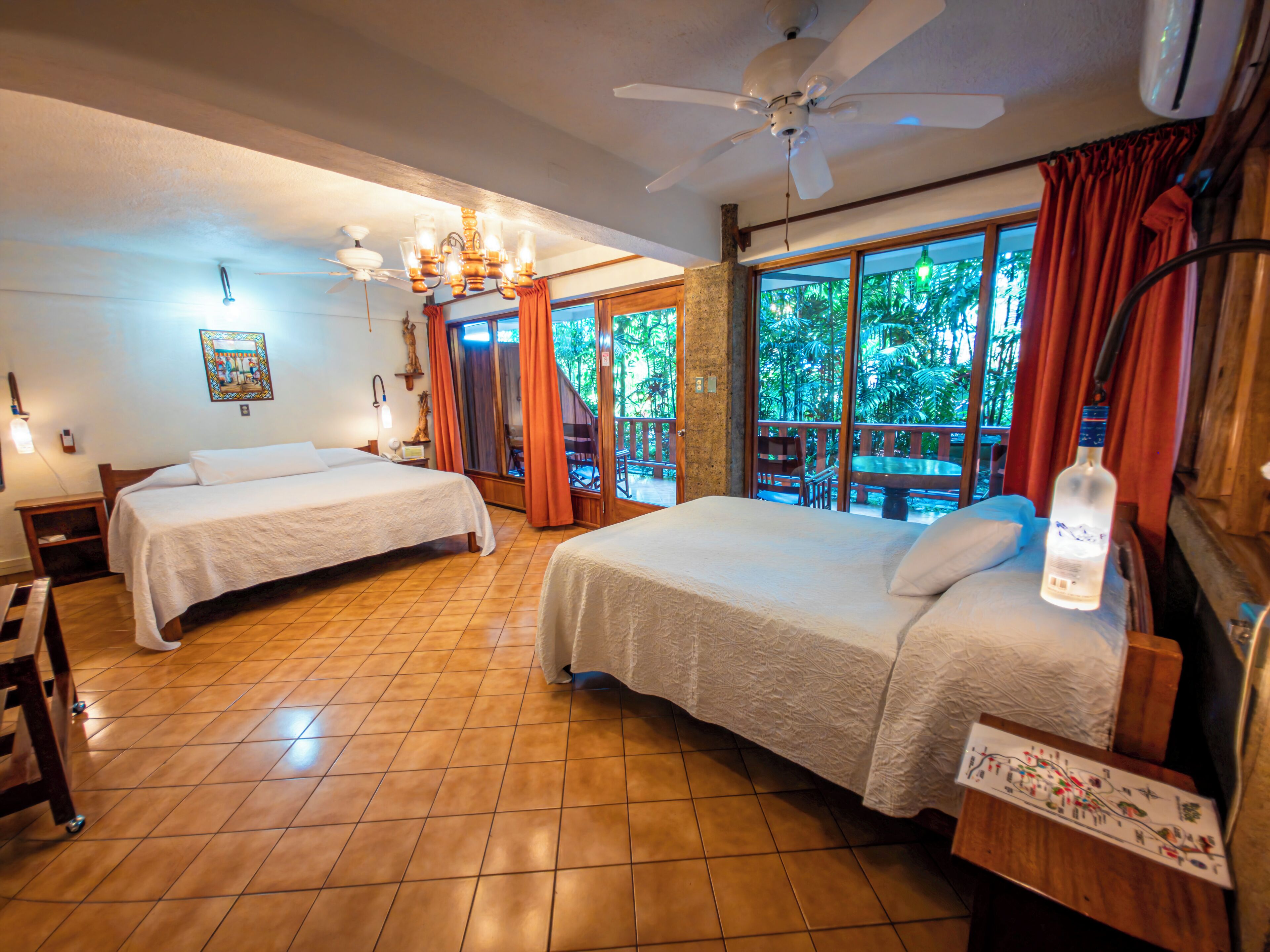 A room with two beds, a ceiling fan, a large window with curtains, and a view of greenery outside.