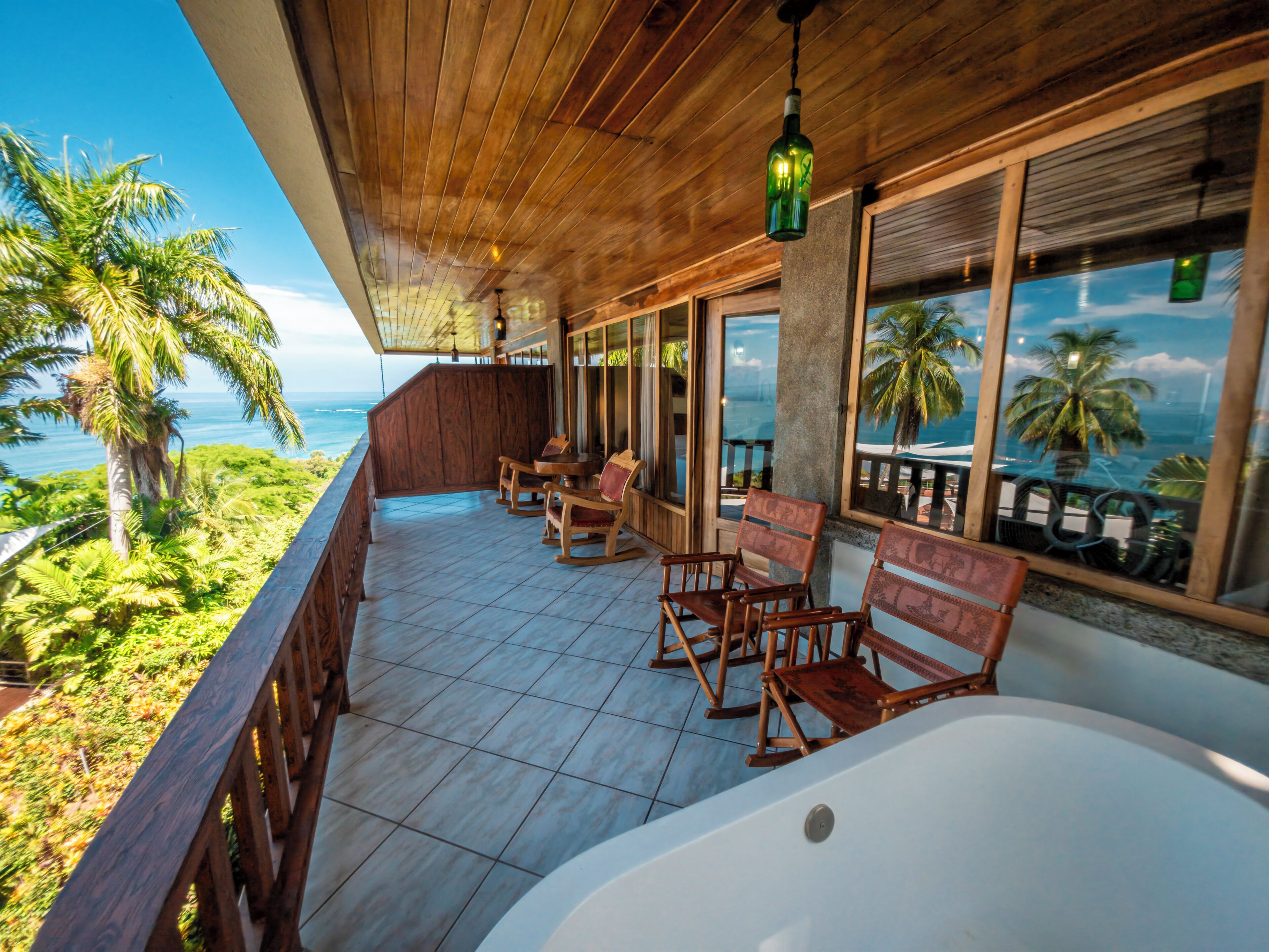 A wooden deck with a view of the ocean, wooden rocking chairs, and a large bathtub.