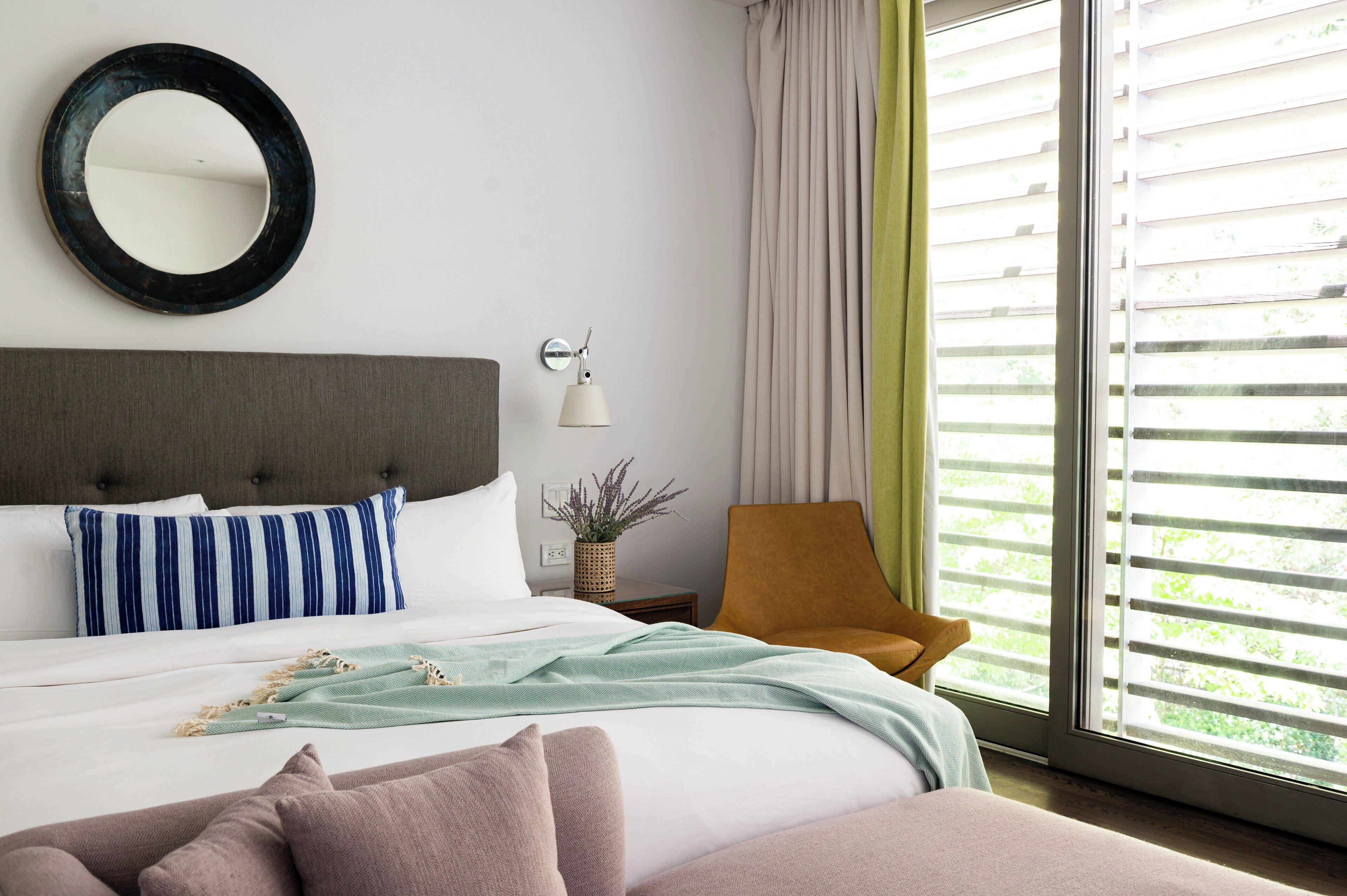 A neatly made bed with a striped pillow, a bedside lamp, and a view of greenery through the window.