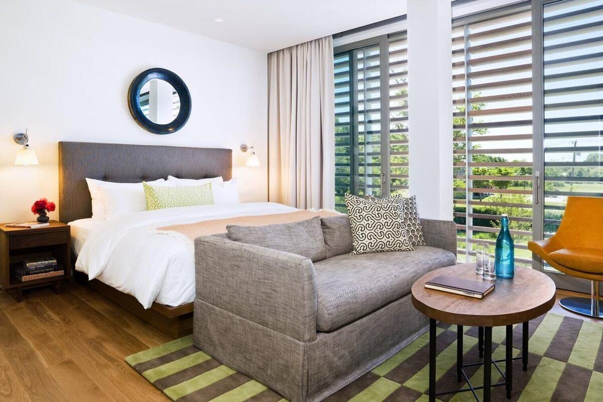 A hotel room with a bed, a sofa, a round table, and a window with blinds.
