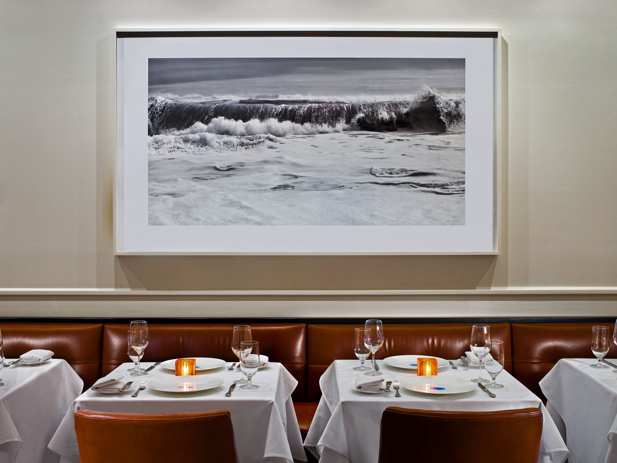 A dining area with tables set for two, each with white tablecloths, wine glasses, and cutlery. A framed photograph of a beach scene hangs on the wall above the tables.