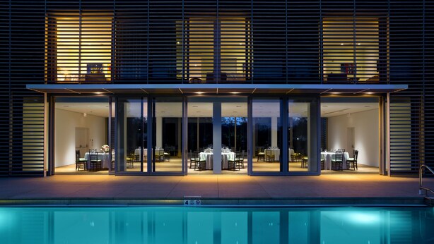 A modern building with a pool, featuring a dining area with tables and chairs, and a glass entrance. - Topping Rose House (Bridgehampton)