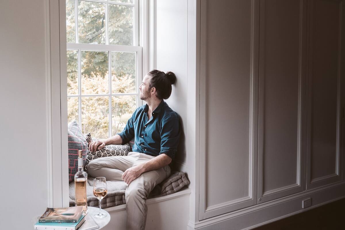 A person sitting by a window in a room with wooden flooring, a small table with a book and a glass, and a pair of shoes on the floor.