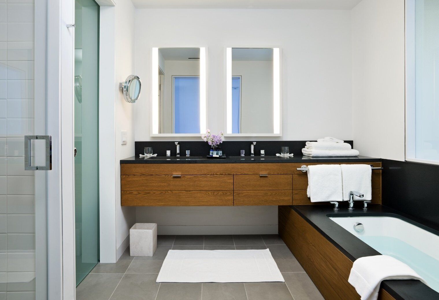 A modern bathroom with a glass shower enclosure, a wooden vanity with a black countertop, a large mirror, and a bathtub.