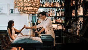 A bar setting with a bartender and a customer. The bar has shelves with various bottles and glasses. There is a cushioned bench in the foreground. - Topping Rose House (Bridgehampton)