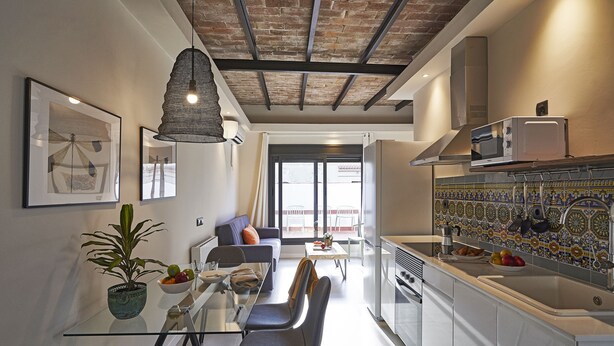 Cocina moderna con microondas, lavaplatos y salpicadero de azulejos. Hay un comedor con mesa de vidrio y sillas, y una sala de estar con sofá y planta.