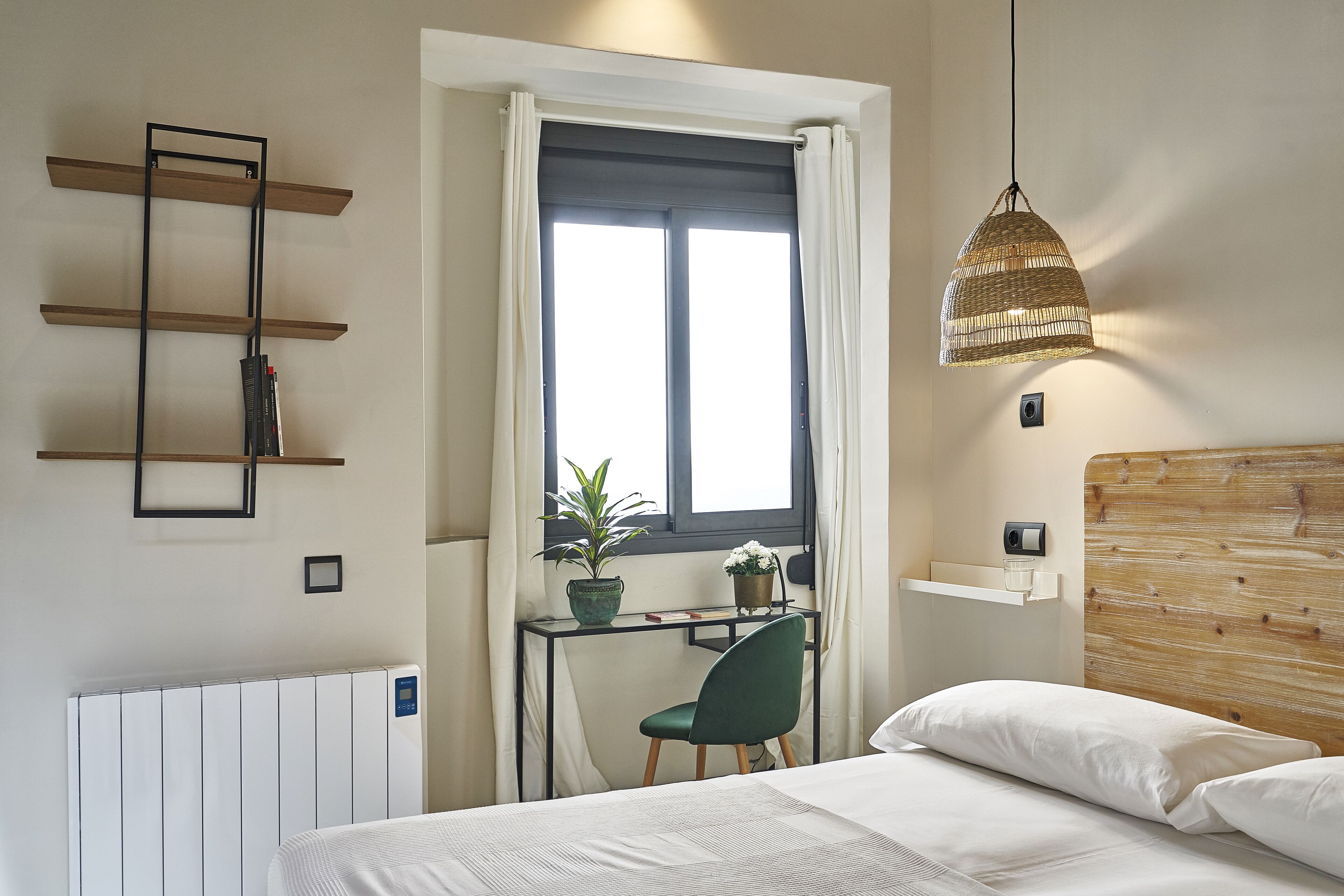 Een slaapkamer met een bed, een bureau met stoel, een raam met gordijnen, een hanglamp en een radiator.