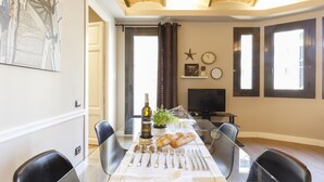 Un comedor con una mesa de vidrio, sillas, una botella de vino, una planta y un libro. Hay ventanas con cortinas, un televisor y un reloj en la pared.