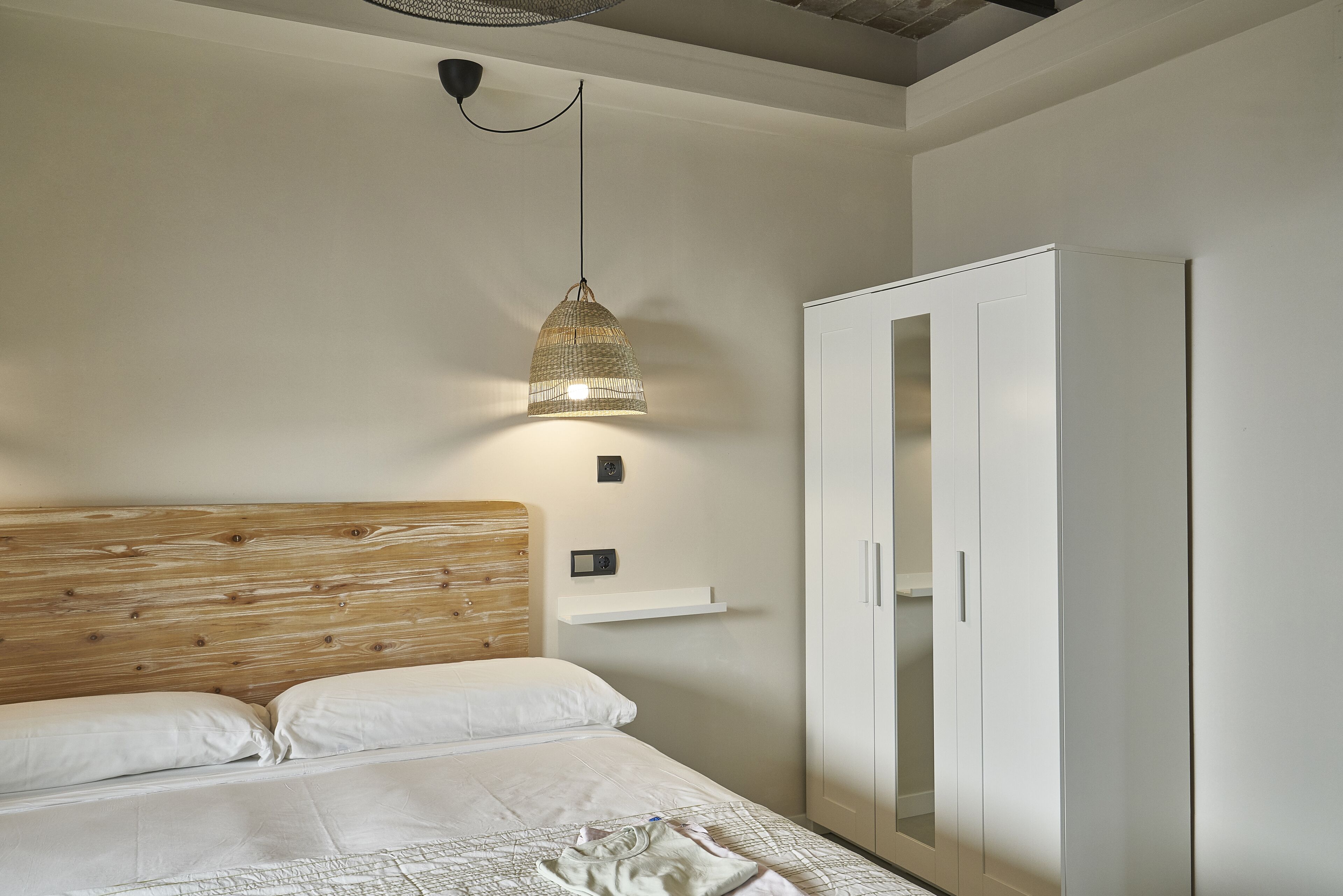 Un dormitorio con cabecera de madera, cama con ropa de cama blanca, lámpara colgante y un armario blanco.