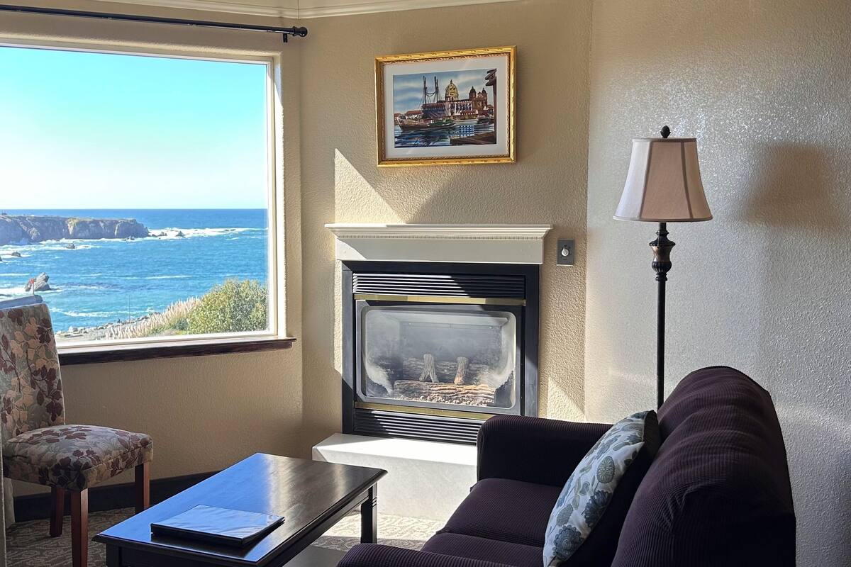 A living room with a fireplace, a sofa, a chair, a table, and a view of the ocean.