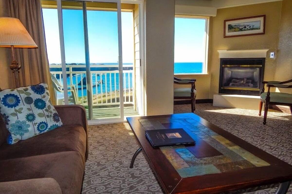 A hotel room with a sofa, a coffee table, a fireplace, and a balcony with a view of the ocean.