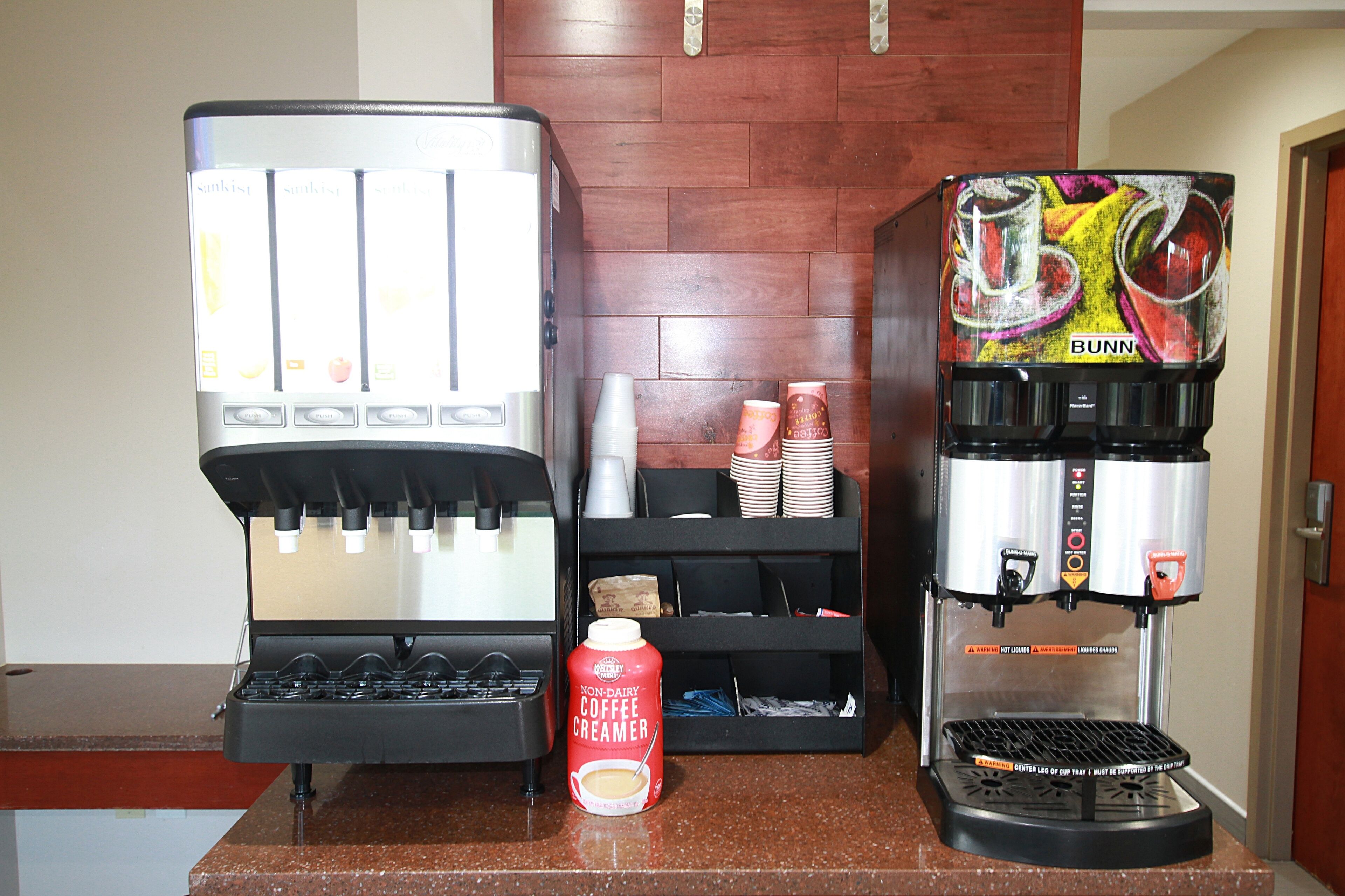 Una estación de café con dos máquinas de café, un dispensador de crema vegetal y vasos desechables apilados.