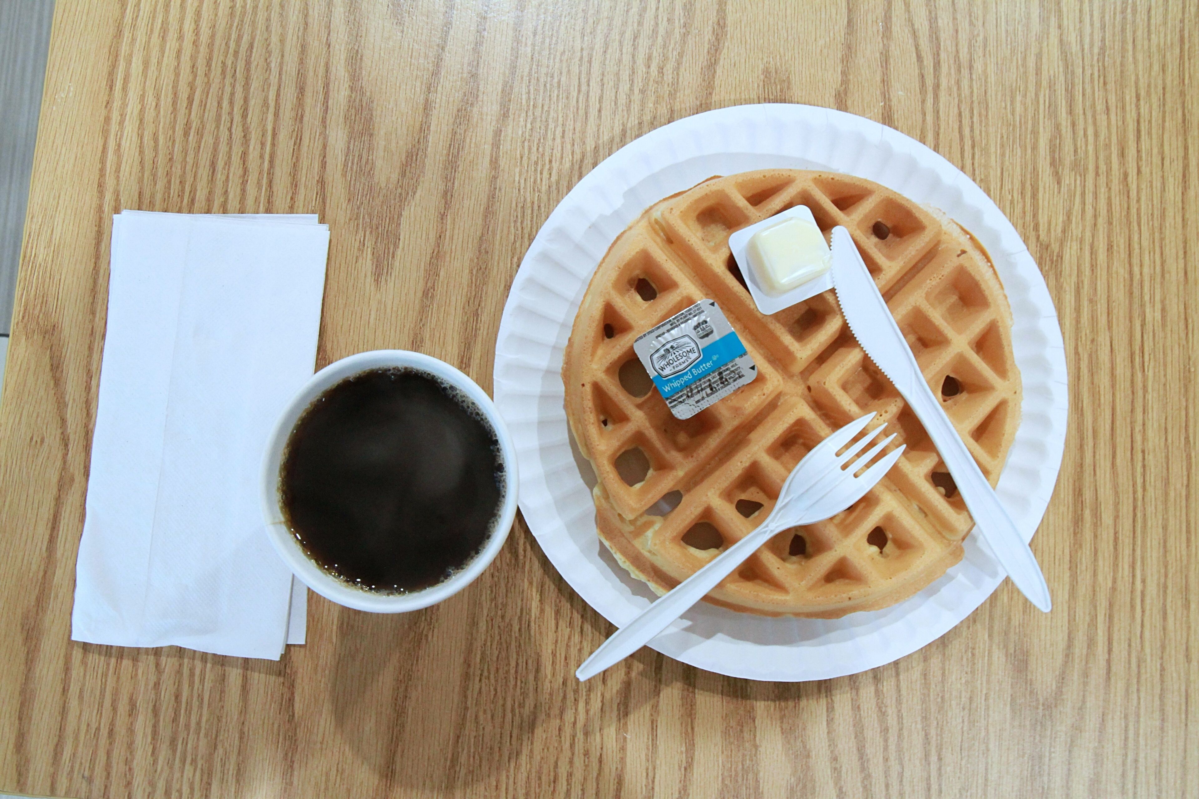 Un waffle con mantequilla y un tenedor sobre un plato de papel, una taza de café y una servilleta en una mesa de madera.
