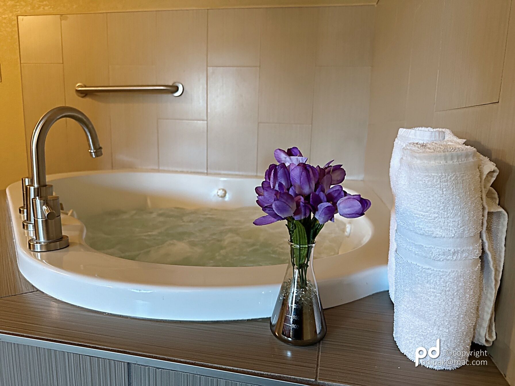 Un baño con una bañera, un grifo, un florero con flores moradas y una pila de toallas blancas.
