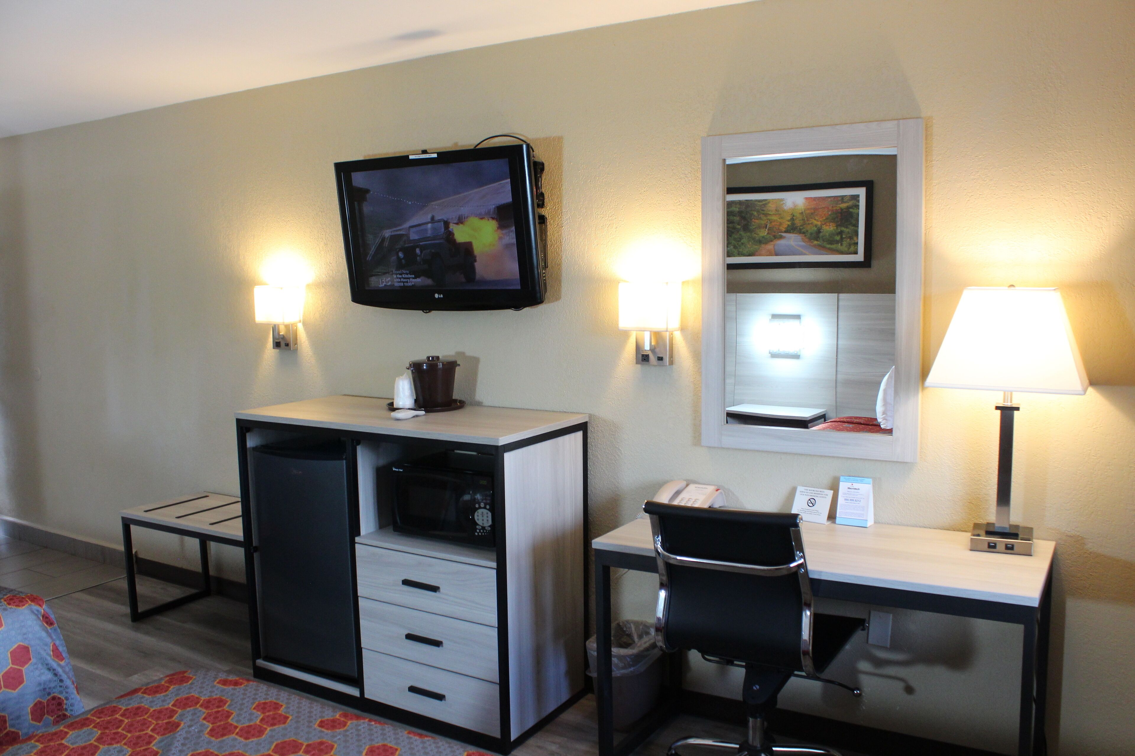 Habitación de hotel con escritorio, silla, televisor en la pared, microondas, cubo de basura y dos lámparas de pared.