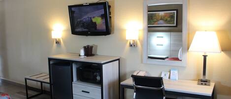 Habitación de hotel con escritorio, silla, televisor en la pared, microondas, cubo de basura y dos lámparas de pared.