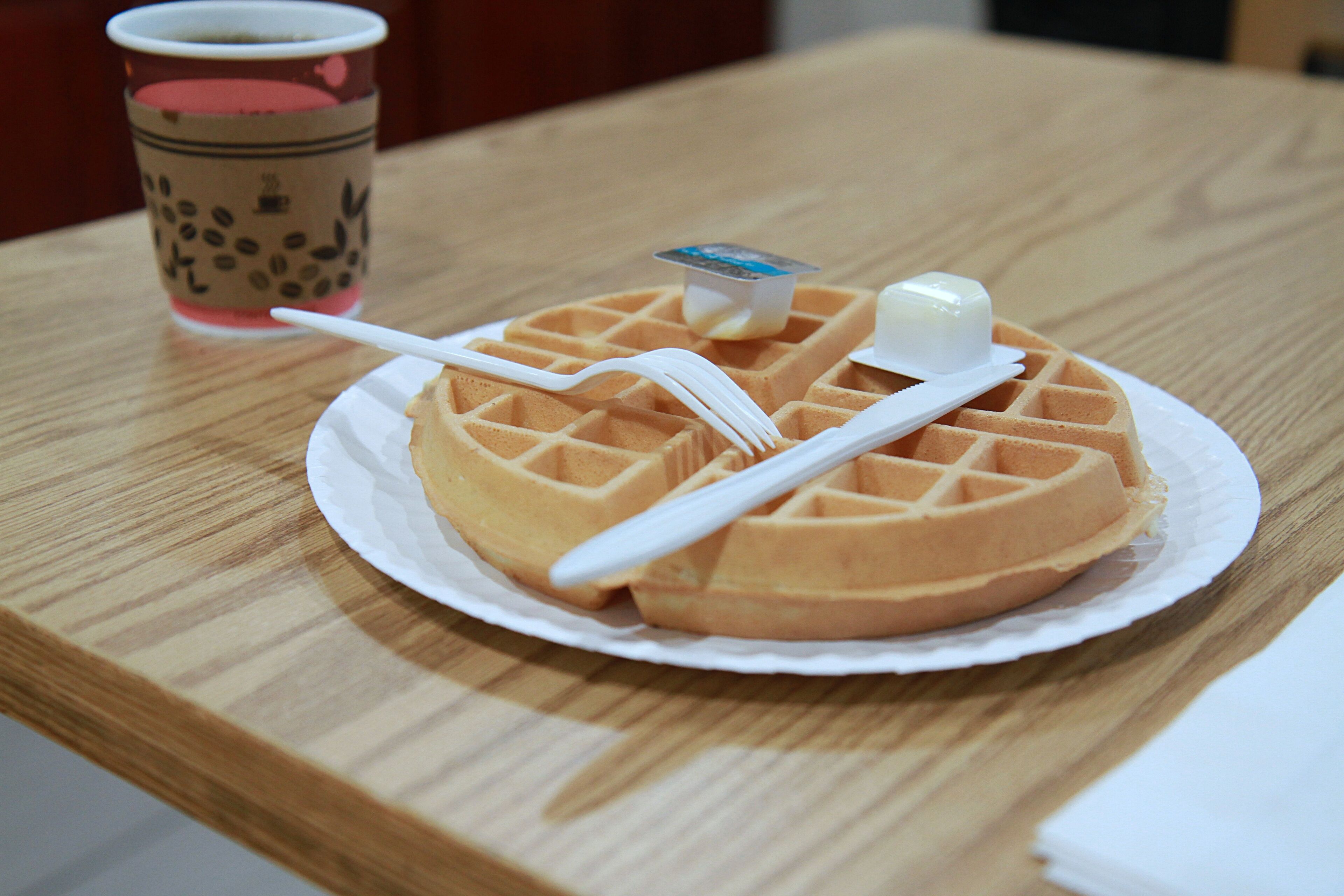 Un waffle con un cuchillo para mantequilla y una cuchara sobre un plato blanco, una taza de té de burbujas y una mesa de madera.