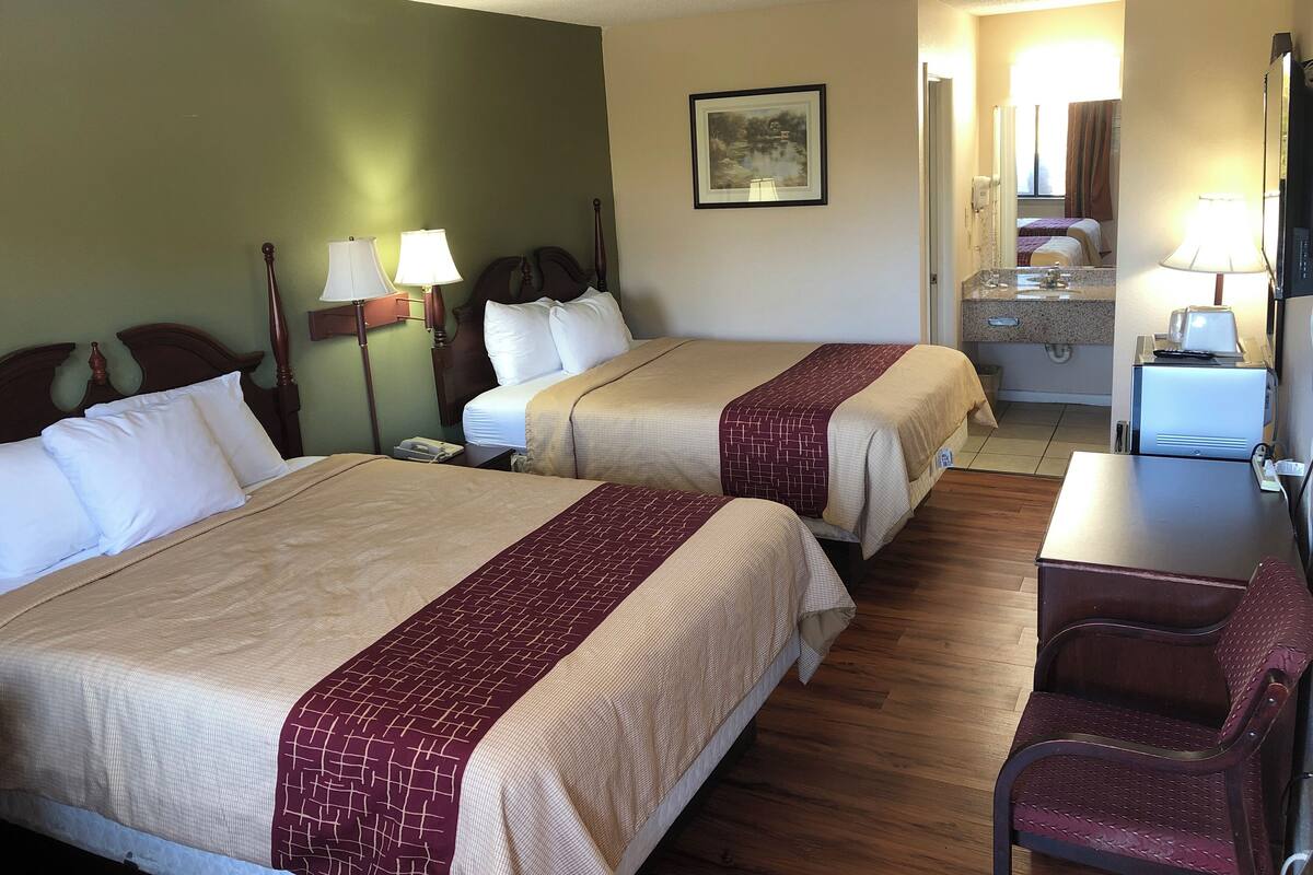 A hotel room with two beds, a desk, a chair, a bathroom, and a painting on the wall.