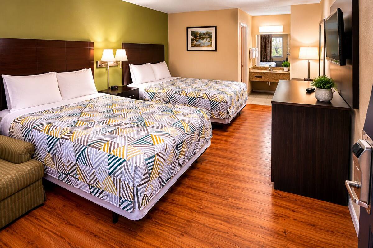A hotel room with two beds, a sofa, a desk, a TV, and a bathroom visible in the background.
