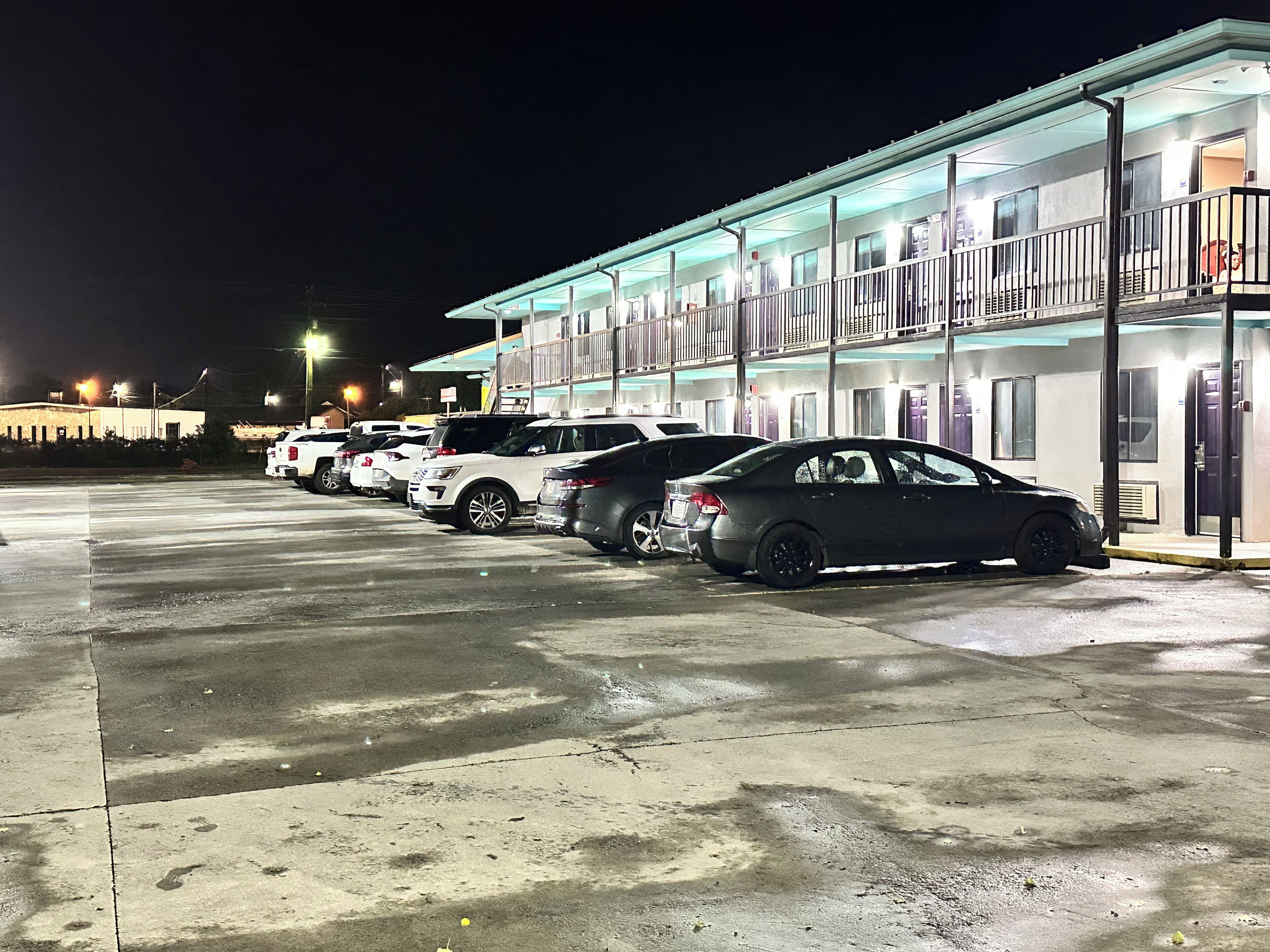 A motel with a row of parked cars, a covered parking area, and a building with multiple windows and doors.
