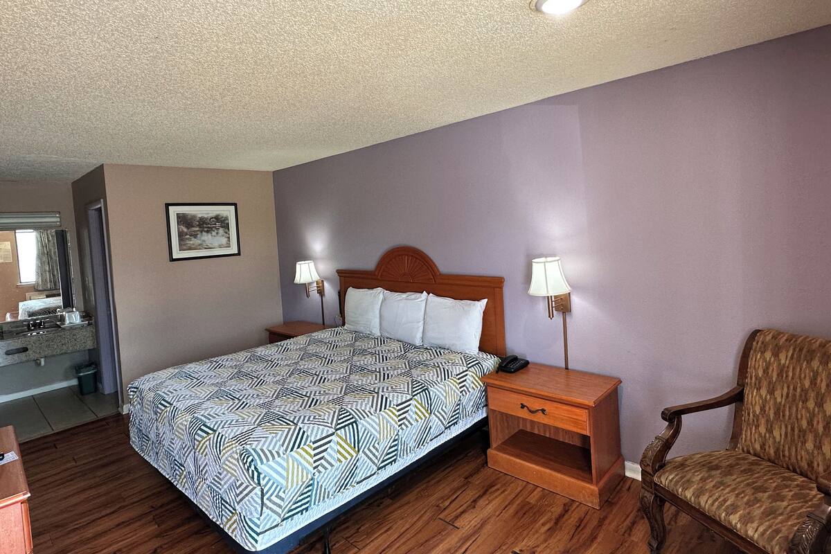 A hotel room with a bed, two bedside tables with lamps, a wooden chair, and a bathroom visible through an open door.