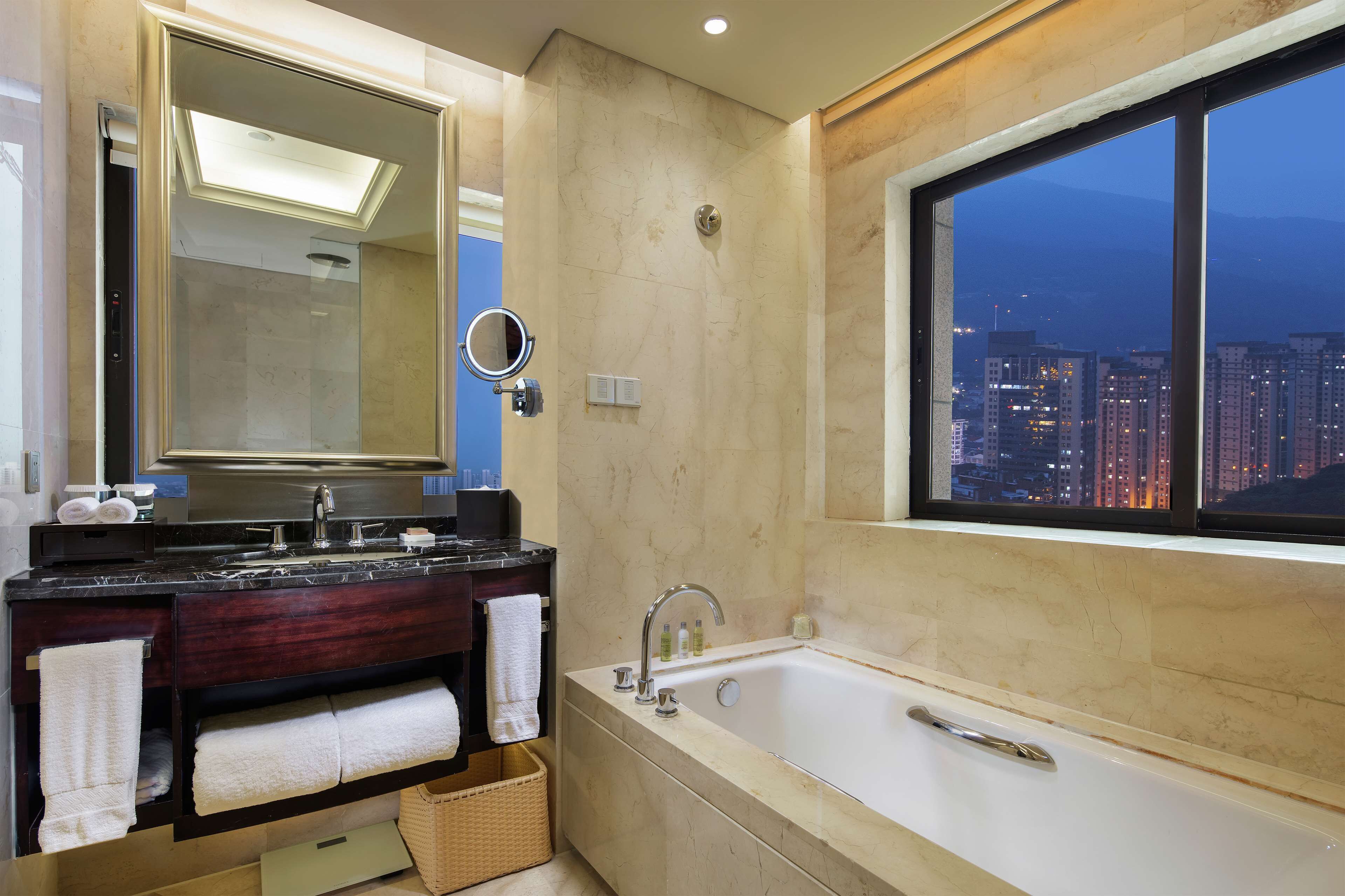 A modern bathroom with a large mirror, a bathtub, and a view of the city at dusk.