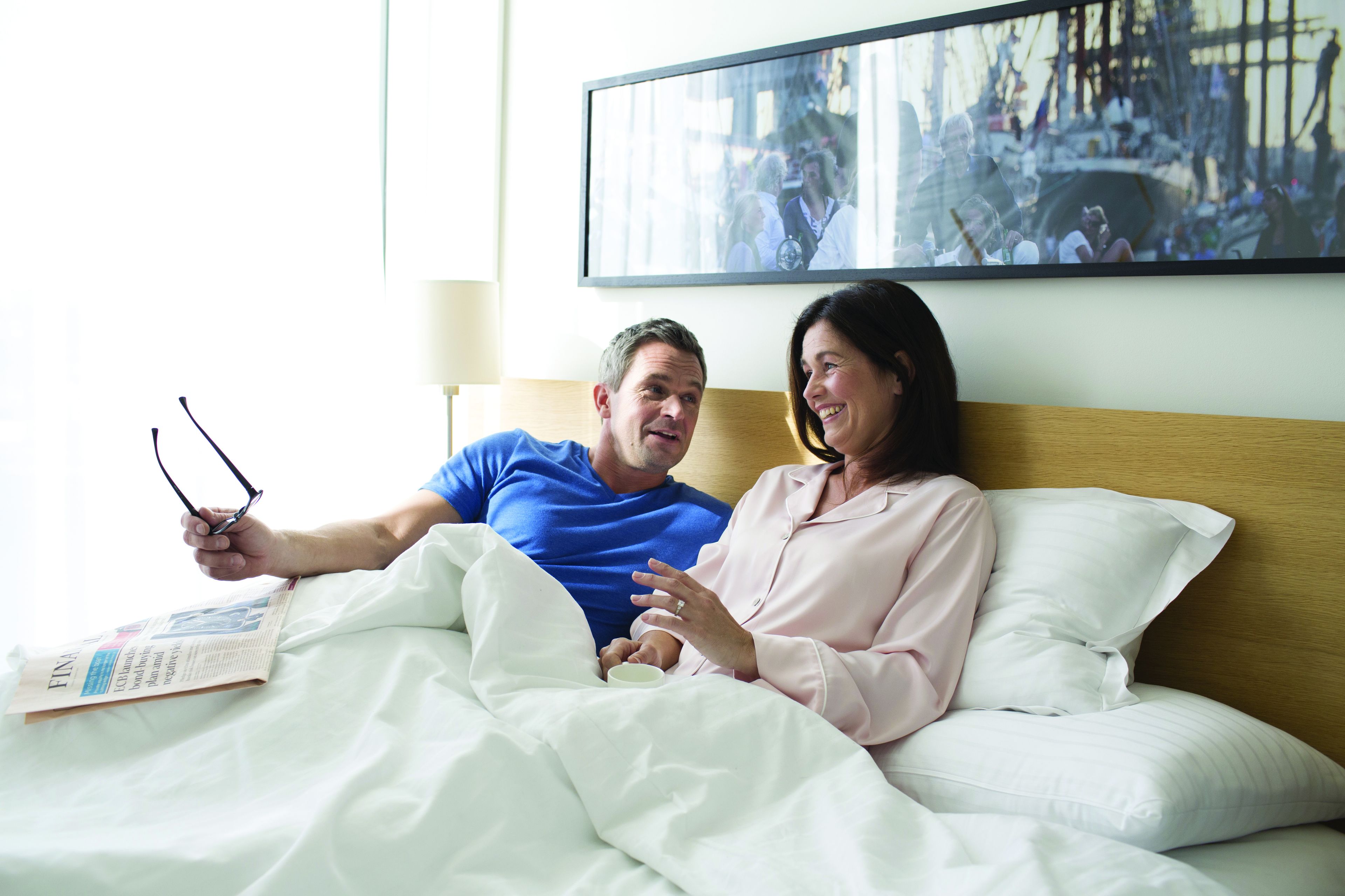 Un couple est allongé dans un lit, un journal est posé à proximité, une photo encadrée est accrochée au mur et une lampe éclaire la pièce.