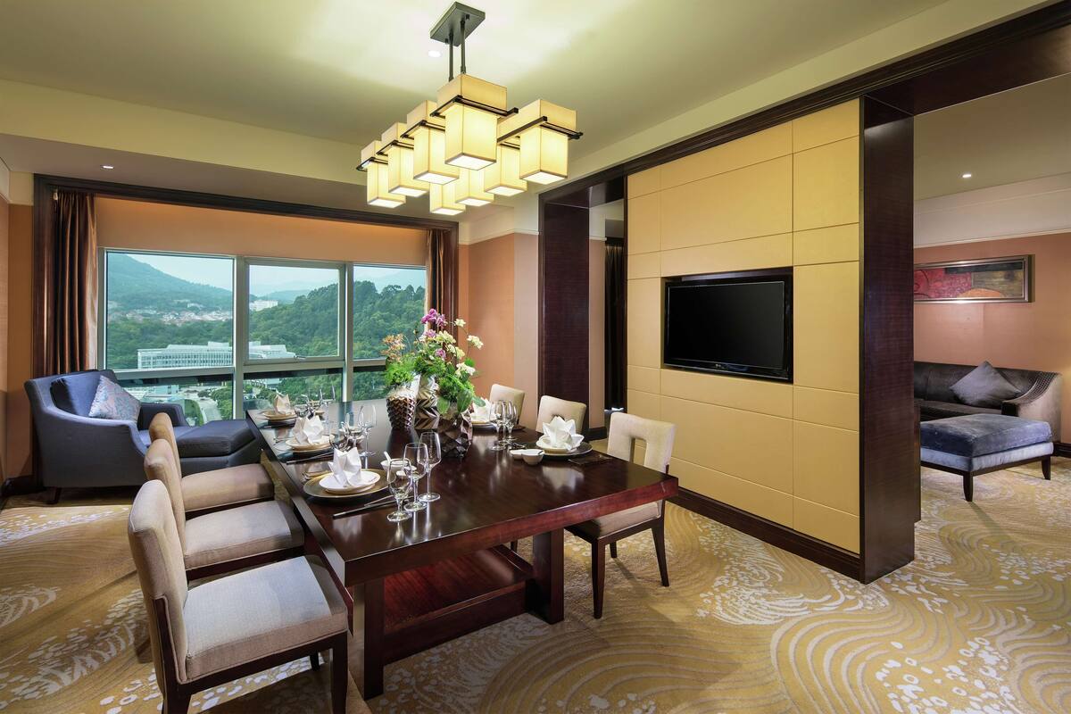 Habitación de hotel con una amplia mesa de comedor para seis personas, un sofá, un televisor montado en la pared y un gran ventanal con vistas al exterior.