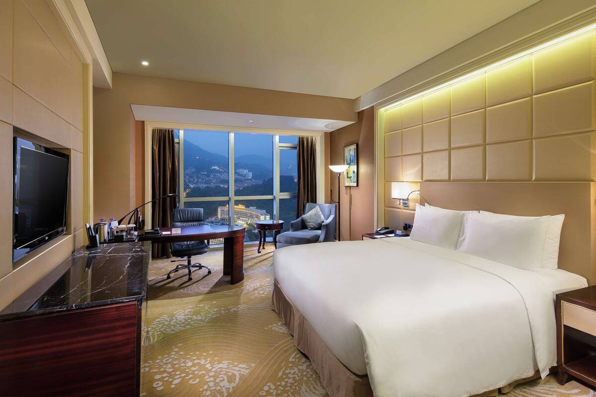 Habitación de hotel con una cama grande, un escritorio, una silla y un televisor. Se ve una ventana con vistas a la ciudad y a las montañas.