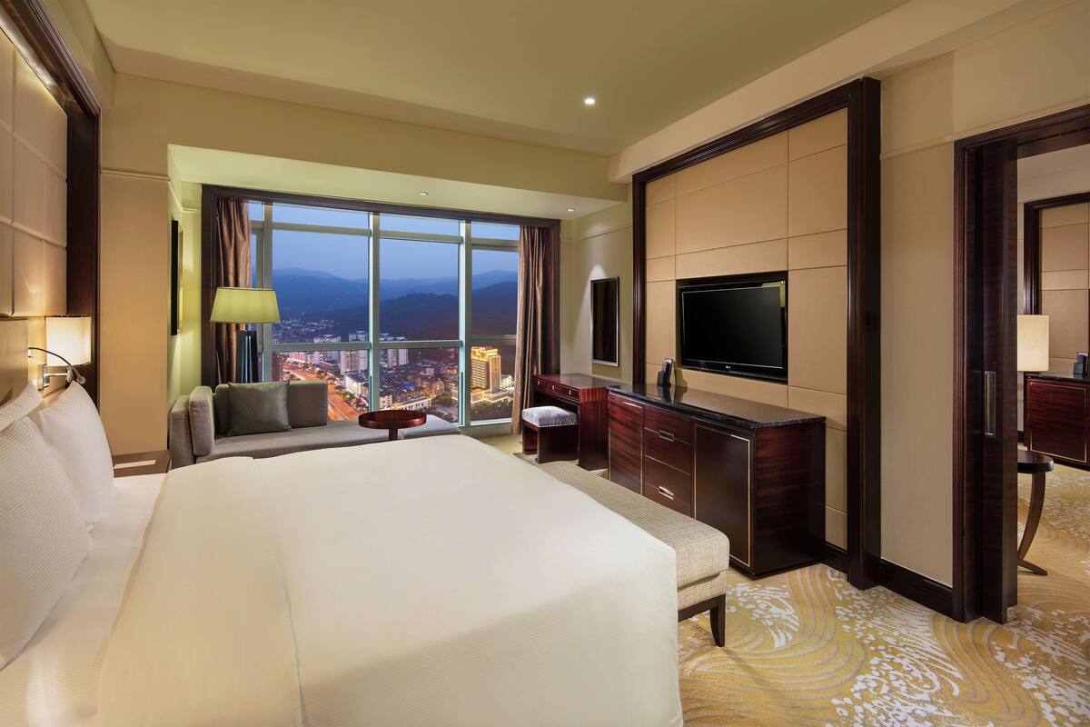 Habitación de hotel con una cama grande, un escritorio, un televisor y vistas a un paisaje urbano a través de un amplio ventanal.