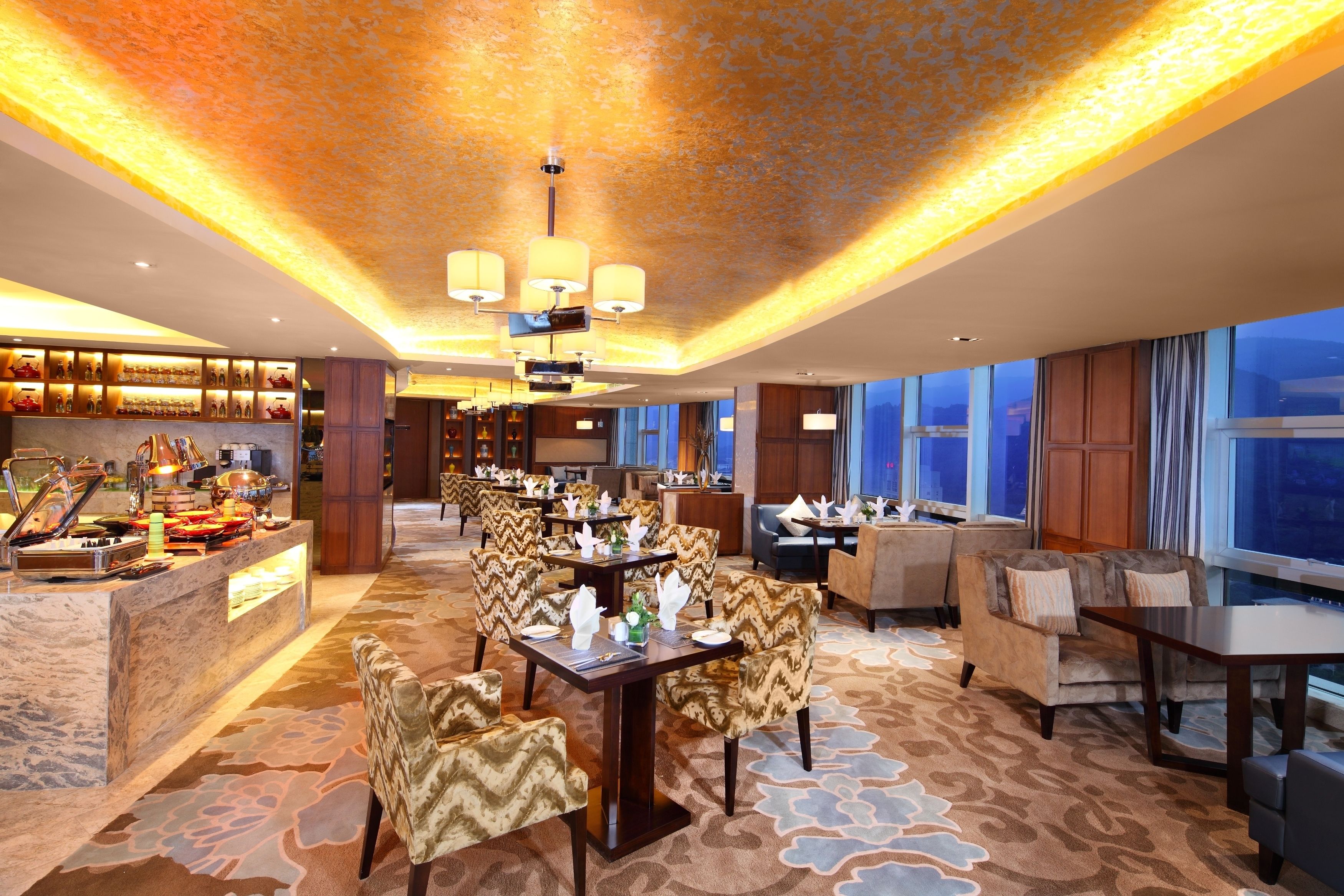Un amplio vestíbulo de hotel con zona de comedor central, mostrador de recepción y grandes ventanales con vistas.
