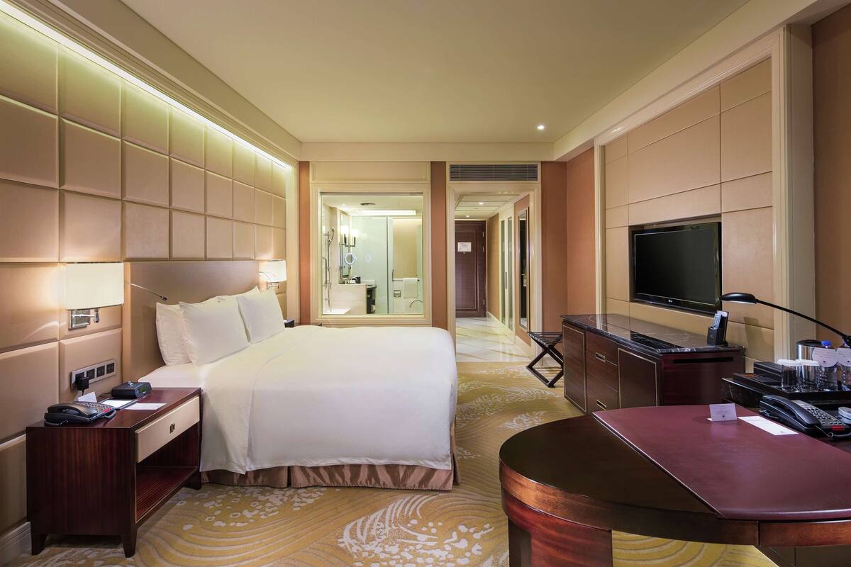 Habitación de hotel con cama, mesita de noche, escritorio, televisor y baño visible a través de una puerta abierta.
