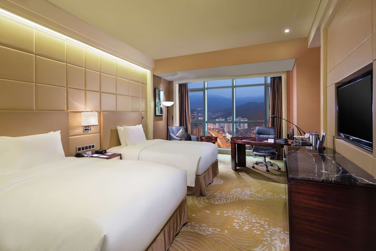 Habitación de hotel con dos camas, un escritorio, un televisor y un amplio ventanal con vistas a la ciudad.