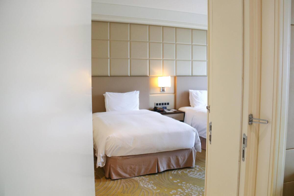 Habitación de hotel con dos camas, cabecera acolchada, lámparas de noche y alfombra estampada.