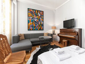 Une chambre d’hôtel moderne avec un canapé gris, une chaise en bois, un lit avec du linge de maison blanc, un tapis noir et blanc, un piano en bois, une télévision et un tableau abstrait coloré accroché au mur.