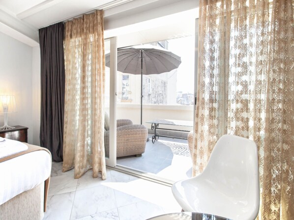 Une chambre d’hôtel avec un lit, une chaise blanche, une porte vitrée donnant sur un balcon équipé d’un canapé en osier et d’un parasol.