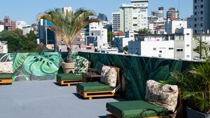A rooftop lounge with wooden loungers and green cushions, a palm tree, and a cityscape view in the background.