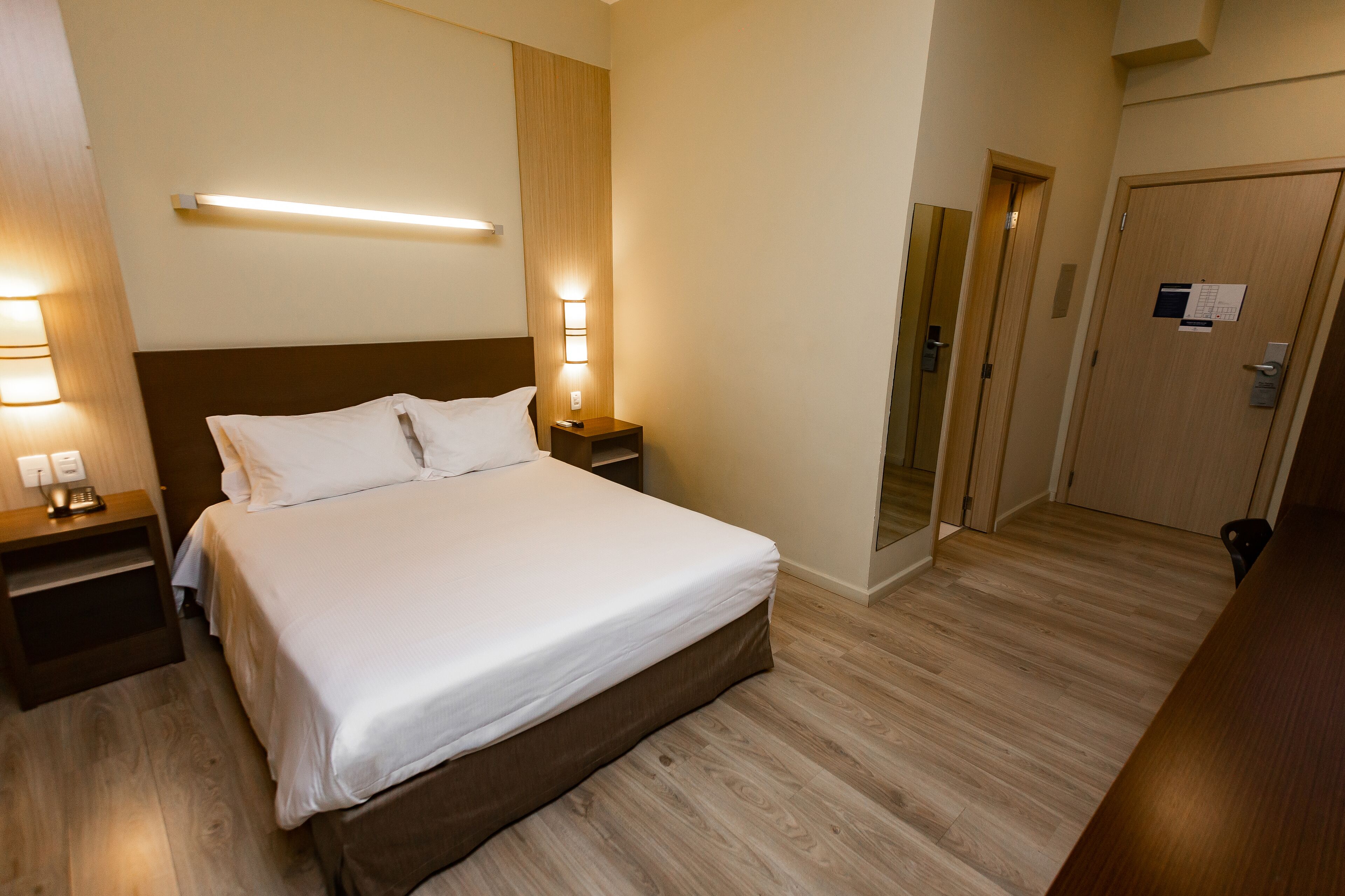 A hotel room with a large bed, wooden flooring, a nightstand with a lamp, and a door with a mirror.