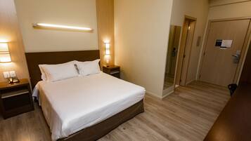 A hotel room with a large bed, wooden flooring, a nightstand with a lamp, and a door with a mirror.