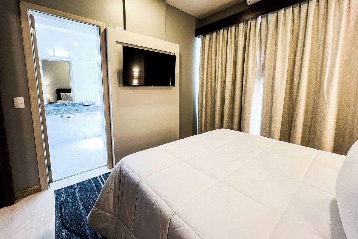 Ein Hotelzimmer mit einem Bett, einem an der Wand befestigten Fernseher, einer Schiebetür zu einem Badezimmer und einem Fenster mit Vorhängen.