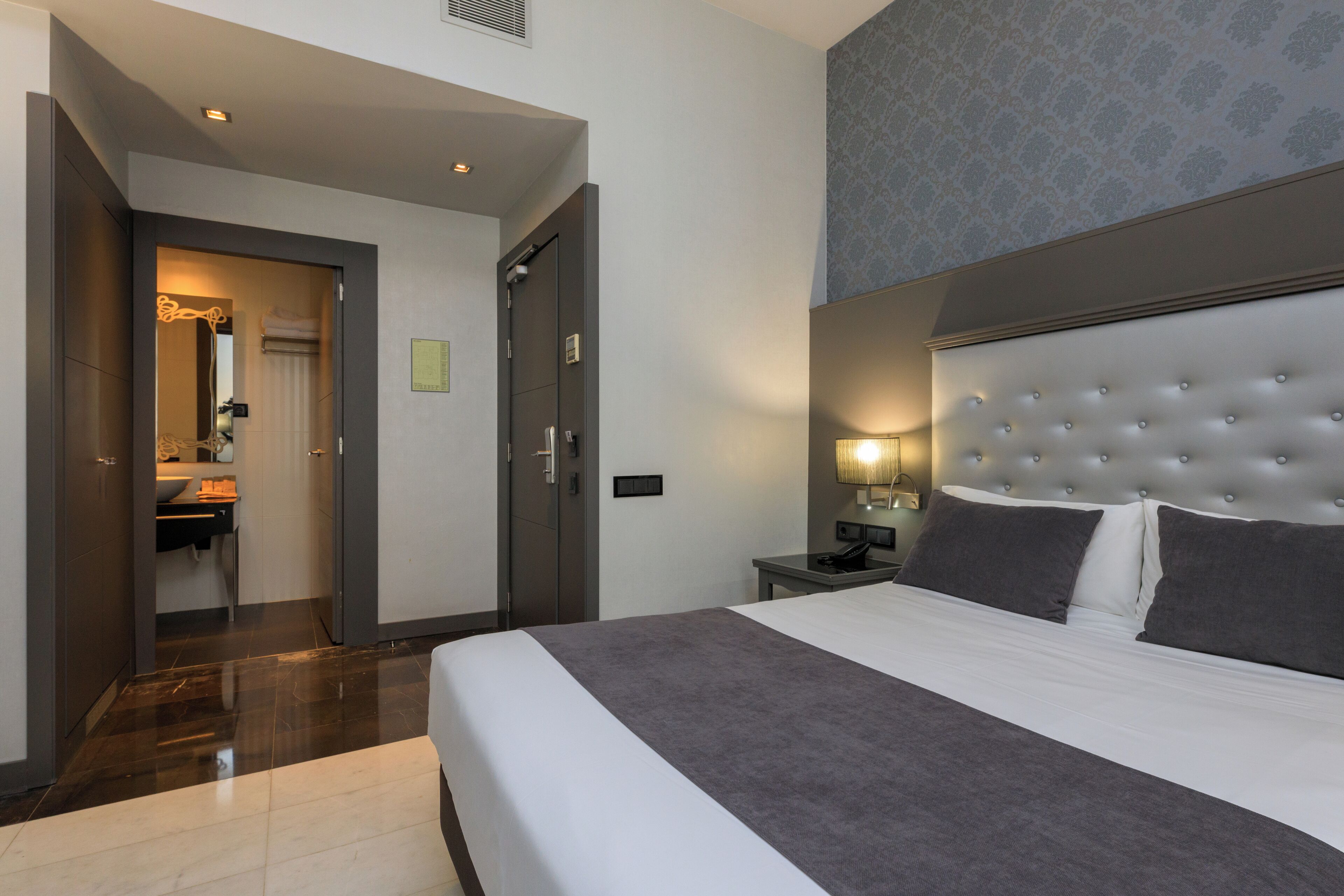 A hotel room with a large bed, a bedside table with a lamp, a wardrobe, and a bathroom visible through an open door.